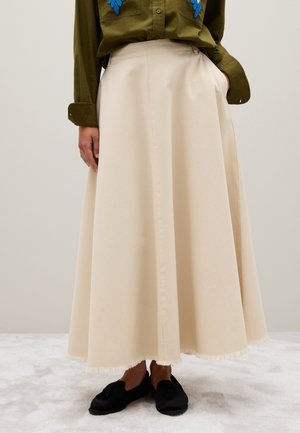 Beige cotton skirt, long and flowing with a fringed hem. Paired with a dark green shirt featuring blue embroidered accents. Black footwear visible.