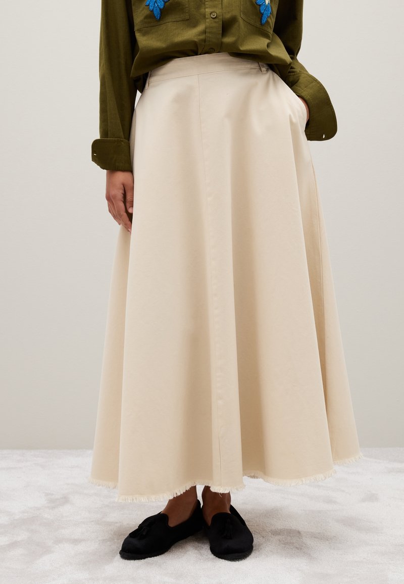 Jupe en coton beige, longue et fluide avec un ourlet à franges. Associée à une chemise vert foncé avec des accents brodés en bleu. Chaussures noires visibles.