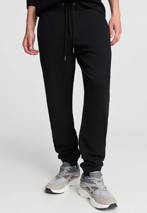 Pantaloni della tuta neri con vita elastica, coulisse regolabile e polsini a zampa, realizzati in tessuto morbido. Indossati con scarpe da ginnastica grigie.