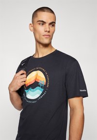 Mørkeblå t-shirt med korte ærmer, der har et rundt grafik af bjerge og træer i orange, gul og teal. Brandlogo på ærmet.