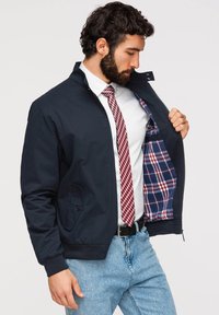 Homme portant une veste bleu marine avec une doublure à carreaux, une chemise blanche, une cravate rouge à carreaux et un jean bleu, montrant l’intérieur de la veste en la tenant ouverte.