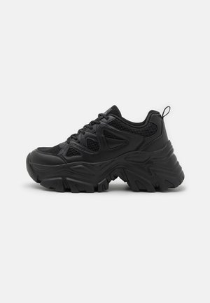 Czarny masywny sneaker z teksturowaną grubą podeszwą, siateczkową i skórzaną cholewką, sznurowaniem z przodu i pętlą na pięcie, widok z boku na białym tle.