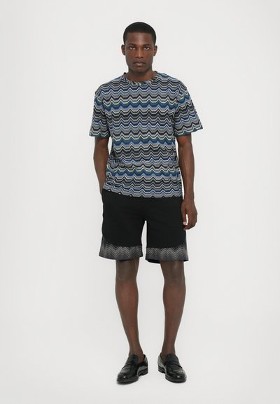 Chemise à manches courtes avec un motif bleu et vert ondulé, associée à un short noir avec une bordure ondulée, complétée par des mocassins noirs.