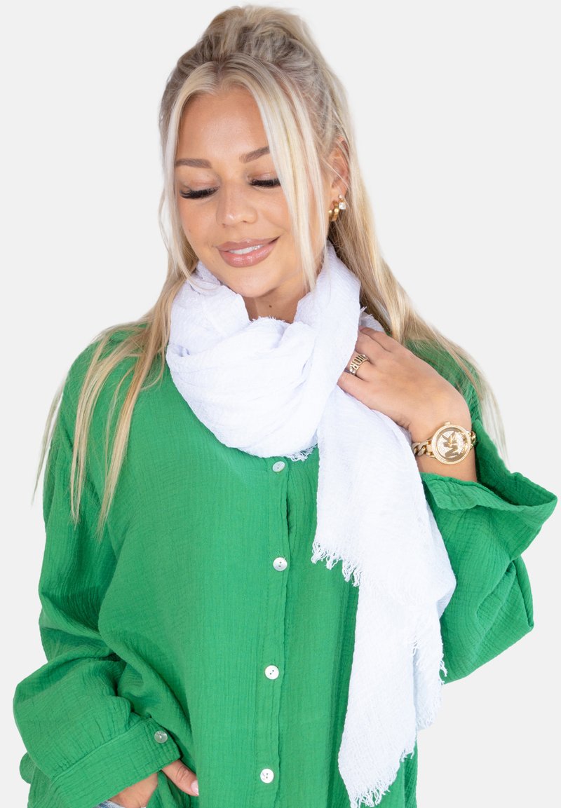 Écharpe blanche texturée drapée autour du cou, contrastant avec une chemise verte à boutons unie. Présente des bords frangés pour un détail supplémentaire.