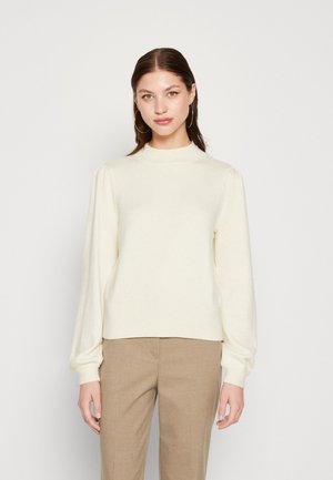 Cream-colored knit sweater with a high neck, dropped shoulders, and ribbed cuffs, paired with light brown trousers.