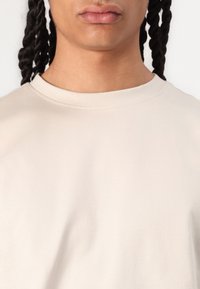 Camiseta de algodón de un beige claro con cuello redondo y mangas cortas. Textura suave y costura sutil a lo largo del cuello.