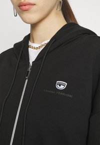 Musta vetoketjullinen huppari, jossa on valkoinen kirjailtu silmälogo ja teksti. Yksityiskohtiin kuuluu säädettävä huppu ja hopeanvärinen vetoketju, jossa on kontrastitikkaukset.