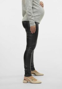 Zwarte faux leren leggings met een slanke pasvorm, gecombineerd met een lichtgrijze trui en beige blokhakken. Het model toont een zijaanzicht.
