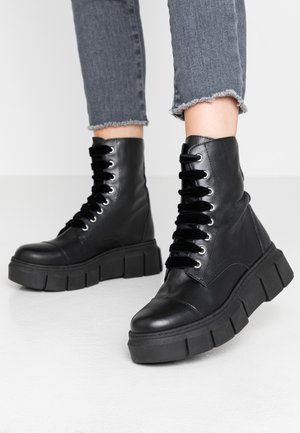 Bottes à plateforme noires lacées portées avec un jean gris raccourci à ourlet effiloché sur un fond clair uni.