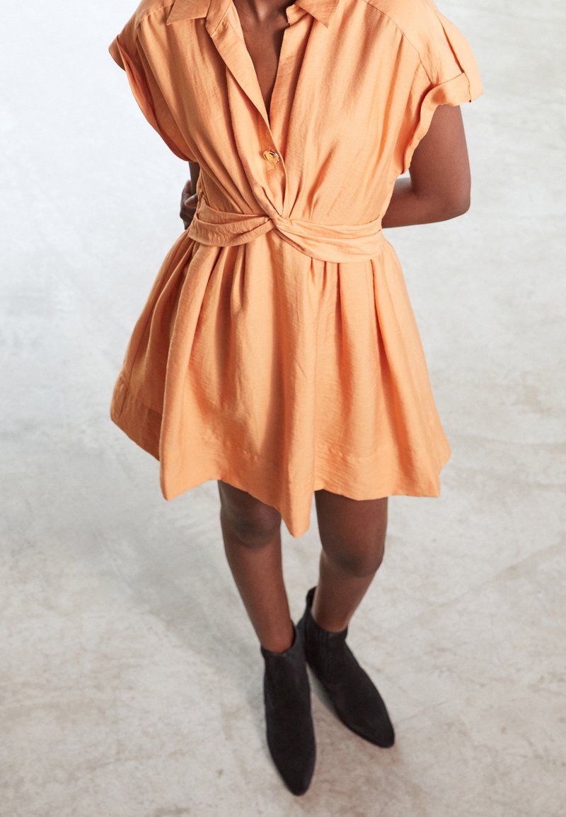 Personne portant une robe pêche à manches courtes avec un nœud à la taille et des bottines noires, debout sur un sol en béton clair.