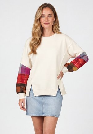 Donna con capelli lunghi e mossi che indossa una felpa crema con maniche a quadri multicolore e una gonna di jeans azzurro chiaro, in piedi con la mano sul fianco.