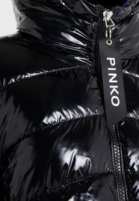 Lśniąca czarna kurtka puchowa zbliżenie na zamek błyskawiczny z etykietą "PINKO" w białym tekście na czarnym tle.
