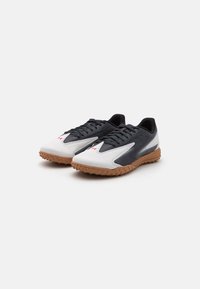 Scarpe sportive con una parte superiore in rete nera con accenti grigi e una punta bianca. La suola è in gomma marrone testurizzata con tacchetti per trazione.