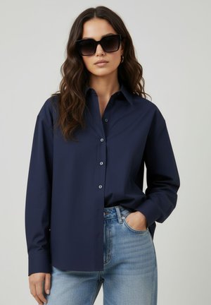 Femme portant une chemise boutonnée bleu foncé oversize, un jean bleu clair, de grandes lunettes de soleil noires et des boucles d'oreilles créoles en or, posant avec une main dans la poche.