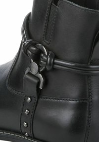 Bottine en cuir noir avec une surface texturée, une boucle en métal ronde et une sangle décorative avec de petites clous en métal sur le côté.