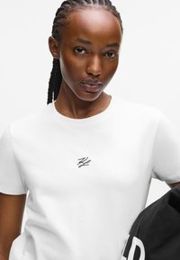 T-shirt bianca in cotone con maniche corte e colletto rotondo, decorata con un piccolo logo ricamato in nero sul petto.