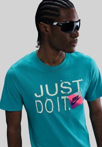 Camiseta de algodón color turquesa con un gran texto blanco que dice "JUST DO IT" y una etiqueta de logo de Nike en rosa. El modelo lleva gafas de sol negras con acentos blancos.
