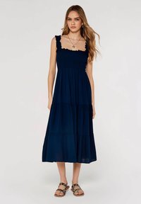 Marineblaues Maxikleid mit ruffled Trägern, gerafftem Oberteil und gestuftem Rockdesign. Kombiniert mit braunen Sandalen mit Nietenakzenten.