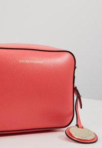 Różowa skórzana torebka o teksturowanej powierzchni, z czarnym wykończeniem i złotymi elementami z logo. Zawiera okrągłą zawieszkę przymocowaną do zamka.