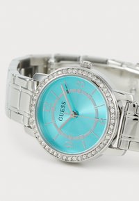 Stålur med en akvablå urskiva, silvriga detaljer och kristalldekorationer runt bezel. Utrustad med en länkad armbandsrem.