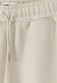 Pantalon de survêtement beige clair avec taille élastique, cordons assortis et une étiquette de vêtement visible glissée à l'intérieur.