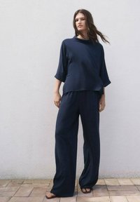 El modelo se encuentra de pie con confianza frente a una pared neutra, vistiendo un atuendo elegante en azul marino que consiste en una blusa suelta y pantalones de pierna ancha, combinados con sandalias.