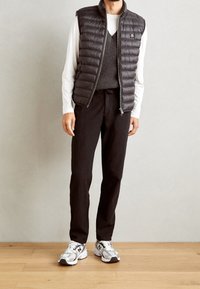 Zwarte puffer vest over een grijze gebreide trui, gecombineerd met zwarte broek en witte sneakers. Soepele textuur, ritssluiting en subtiele logo-details.