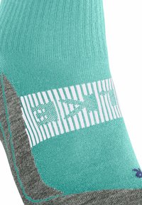 Zbliżenie na turkusową skarpetę z paskiem w kolorze białym i wzorami, kontrastującą z szarym piętą i palcami.