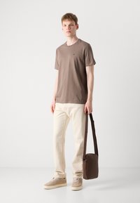 Braunes Baumwoll-T-Shirt, cremefarbene Hose, beige Sneakers und eine braune Umhängetasche mit langem Riemen, stehend vor einem schlichten Hintergrund.