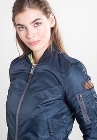 Granatowa bomberka z błyszczącego nylonu. Posiada przedni zamek błyskawiczny, ściągacz przy kołnierzu oraz kieszeń na rękawie zapinaną na zamek z brązową metką.