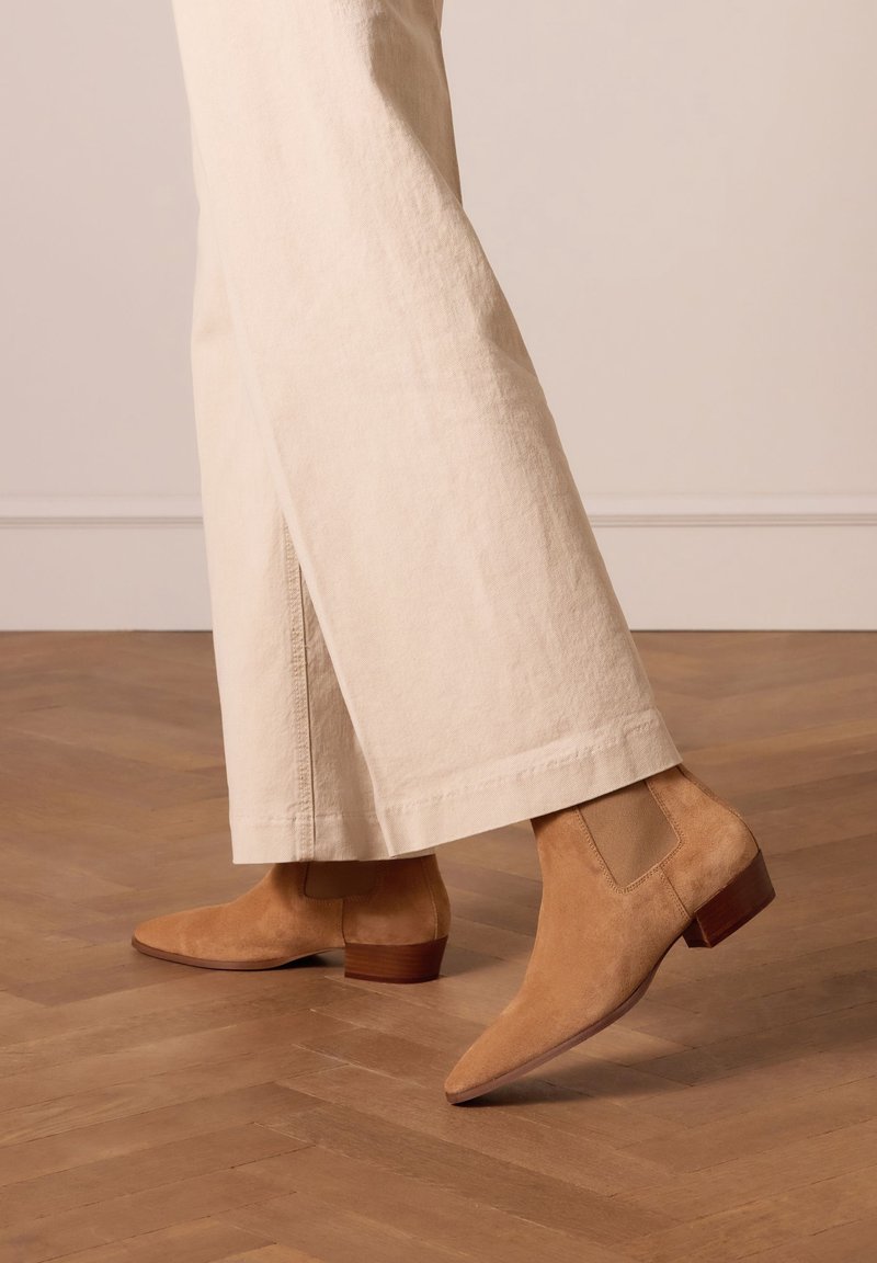 Personne portant un pantalon beige à jambes larges avec des bottines en daim couleur noisette marchant sur un sol en bois à l'intérieur.