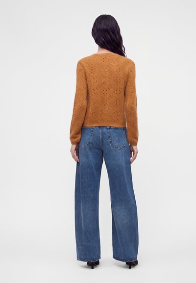 Femme aux longs cheveux foncés portant un pull marron texturé et un jean bleu à jambes larges, debout dos à un fond uni.