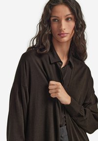 Brun cardigan i mjukt tyg, med lös passform, öppen fram och långa ärmar. Bärs över en mörk skjorta, vilket framhäver texturen.