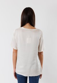 Blusa a maniche corte di colore crema realizzata in un tessuto leggero e liscio, con un taglio rilassato e scollo rotondo, presenta un orlo leggermente curvato.