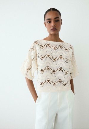 Donna che indossa un top lavorato a crochet largo color crema con motivi ondulati e pantaloni bianchi a vita alta, in piedi su uno sfondo bianco semplice.