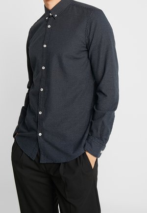 Homme portant une chemise bleu foncé à manches longues avec de petits points blancs et un pantalon noir, les mains dans les poches.