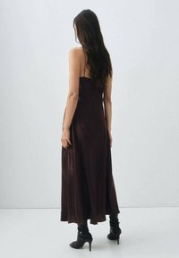 Langes, dunkelbraunes Satin-Kleid mit Spaghettiträgern, das ein fließendes Design und eine glatte Textur aufweist. Komplementiert mit schwarzen Knöchelstiefeln.