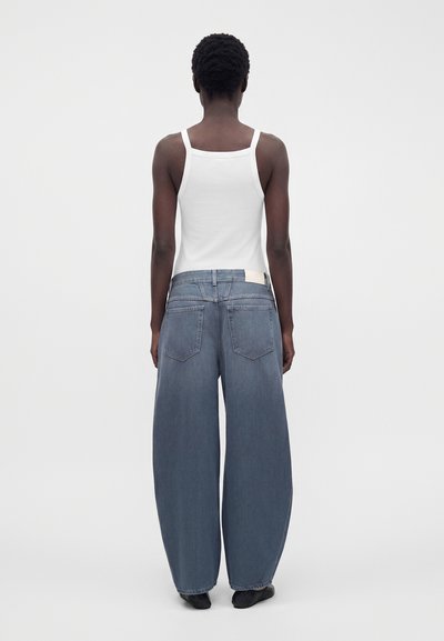 Personne debout de dos portant un débardeur blanc et un jean bleu à jambes larges avec des chaussures noires sur un fond uni.