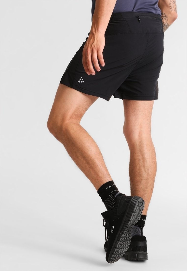 Man som bär svarta träningsshorts, svarta strumpor och svarta löparskor, stående med ett ben korsat bakom det andra mot en vit bakgrund.