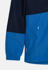 Blauer und dunkelblauer Windbreaker mit lockerem Schnitt, elastischen Bündchen und einer diagonalen Nahtdetails sowie einem kleinen Logo-Akzent.