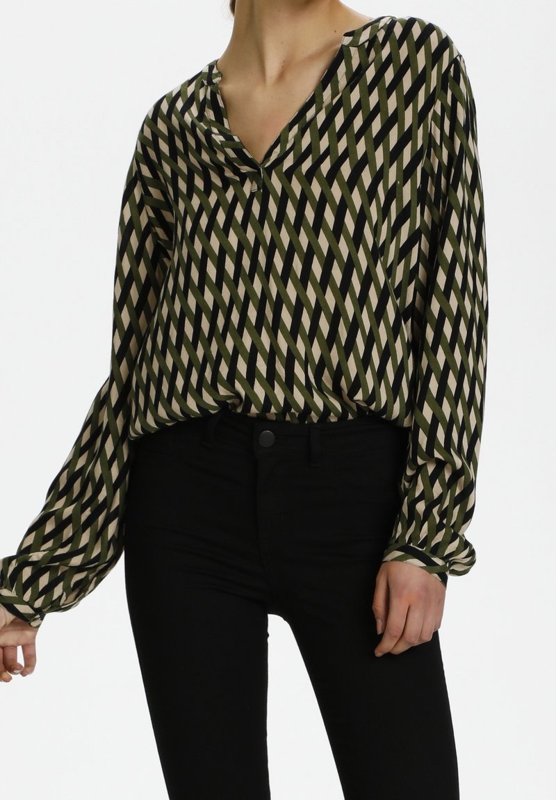 Personne portant un chemisier à manches longues et col en V avec un motif à rayures diagonales vert, noir et beige ainsi qu’un pantalon noir ajusté.