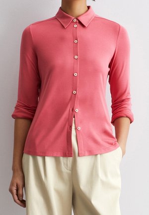 Femme portant une chemise corail à manches longues boutonnée avec les manches retroussées et un pantalon couleur crème, debout contre un mur clair.