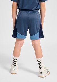 Blå idrottsshorts med mörkblå detaljer, elastisk midja och fickor. Vita strumpor med svart chevronmönster och ljusa skor.