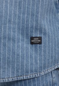 Ανοιχτό μπλε denim ύφασμα με ρίγες και μαύρη ορθογώνια ετικέτα που γράφει "MADS NØRGAARD COPENHAGEN", ραμμένη κοντά στο κέντρο.