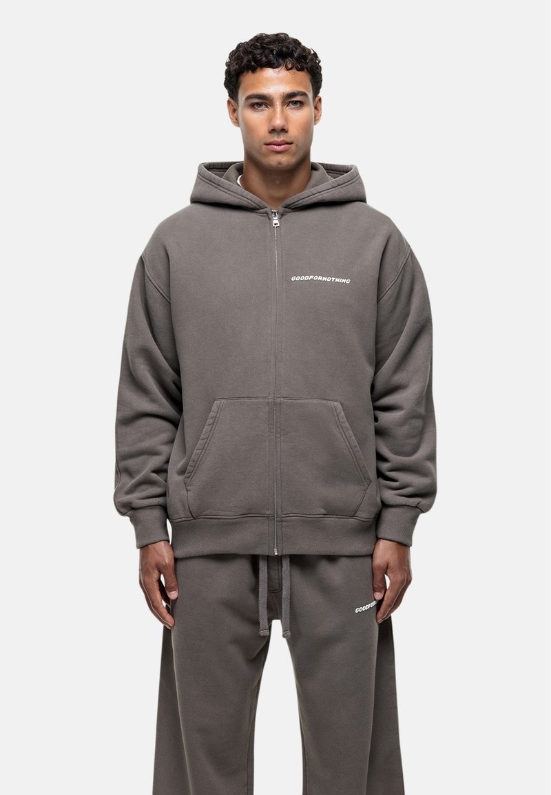 Grauer Reißverschluss-Hoodie aus weichem Stoff, mit einer Fronttasche und weißem Logo-Detail auf der Brust, kombiniert mit passenden Hosen.