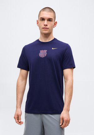 Hombre con cabello corto que lleva una camiseta azul marino con un logo de escudo rojo y azul desvanecido y una pequeña marca amarilla de Nike.