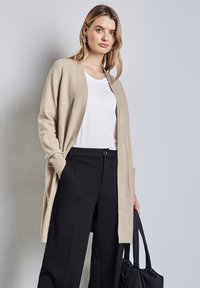 Beige cardigan van zacht materiaal, met een open voorkant, lange mouwen en zijzakken, gedragen over een witte t-shirt en zwarte wijde broek.