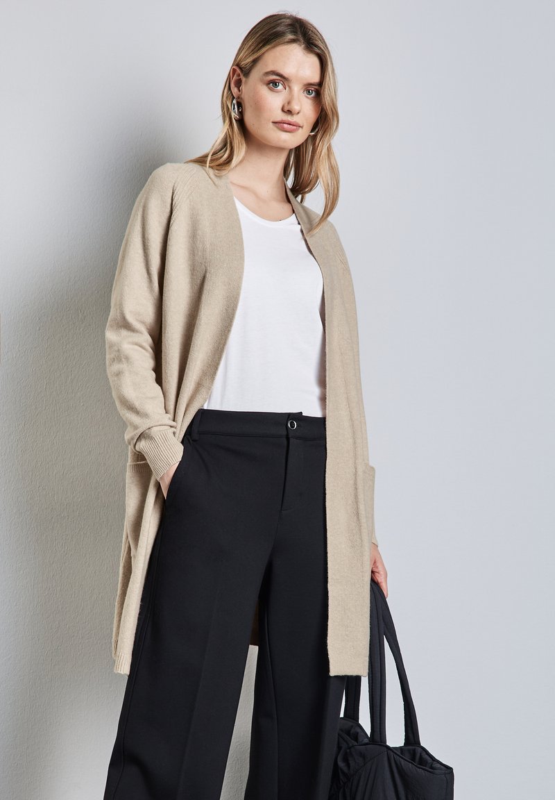 Beige cardigan van zacht materiaal, met een open voorkant, lange mouwen en zijzakken, gedragen over een witte t-shirt en zwarte wijde broek.