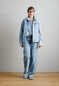 Lichtblauwe denimjas met grote zakken, lichtblauw shirt, wijde jeans en blauwe instappers, gepresenteerd tegen een neutrale achtergrond.