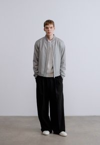 Szürke bomber dzseki fehér bordázott pulóverrel, fekete bő szárú nadrággal és fehér tornacipővel párosítva. Minimalista stílus tiszta környezetben.